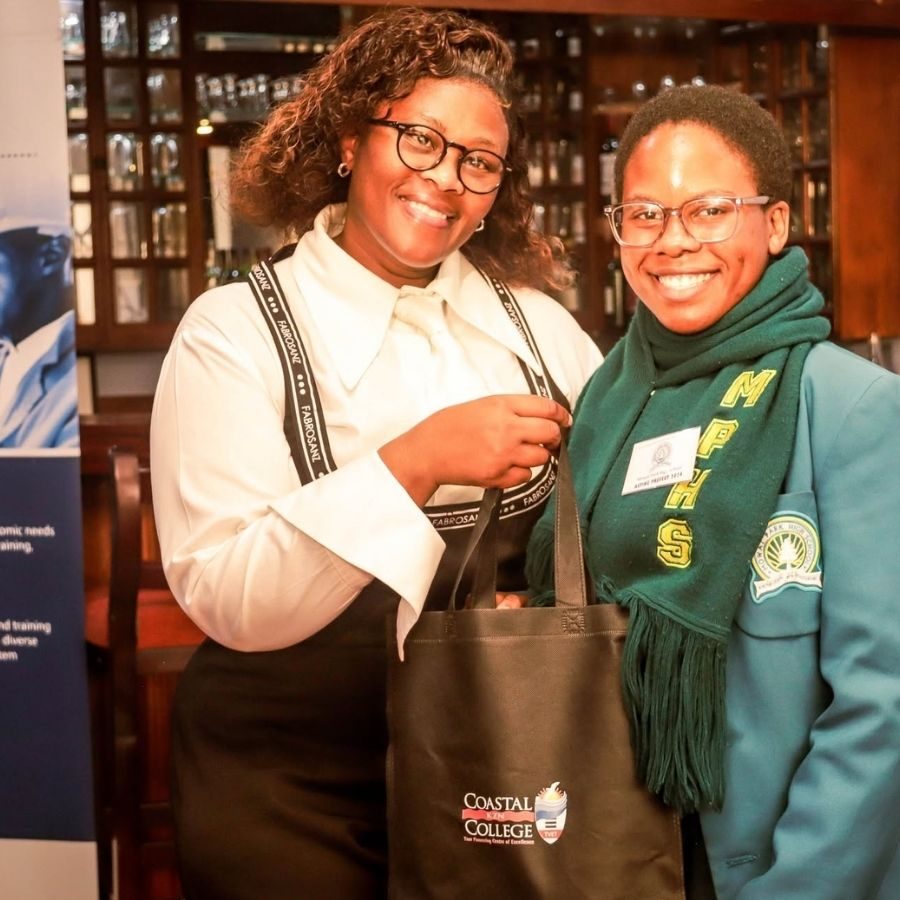 Two smiling students holding a Coastal KZN College tote bag at a college function or awards evening