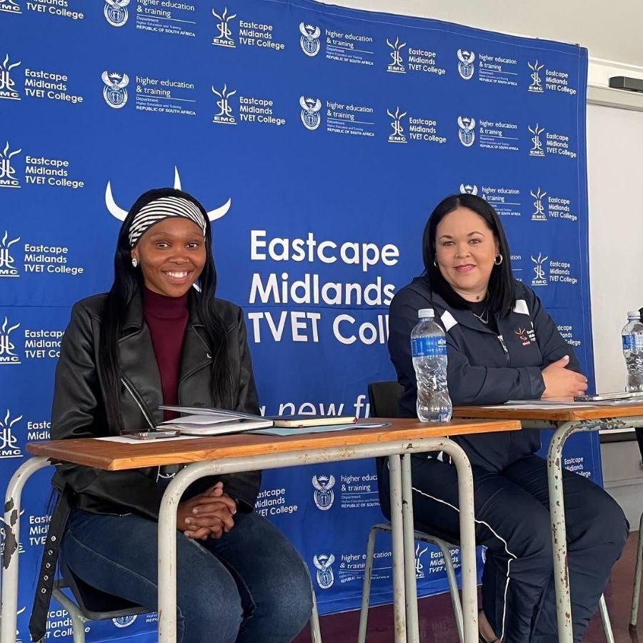 Two Guests Seated at Eastcape Midlands College Event Two women seated at desks in front of an Eastcape Midlands TVET College branded backdrop wall