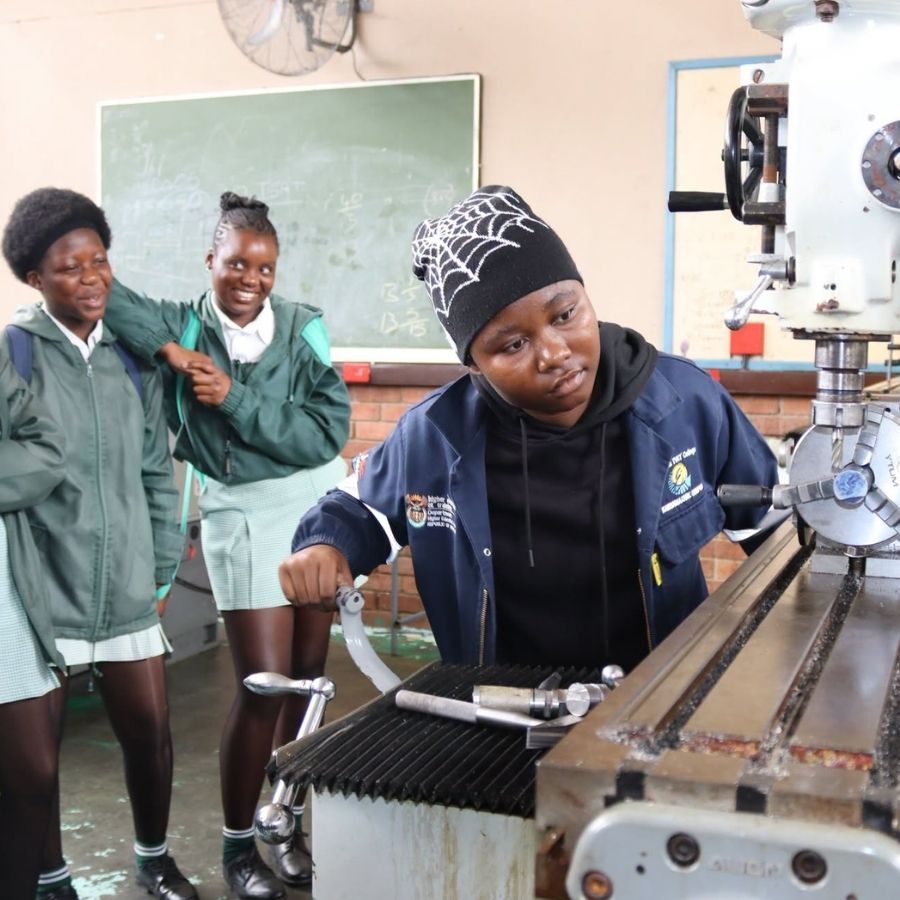 Student Operating Machinery in Engineering Workshop Ehlanzeni TVET College student operating a drill press in an engineering workshop as classmates look on