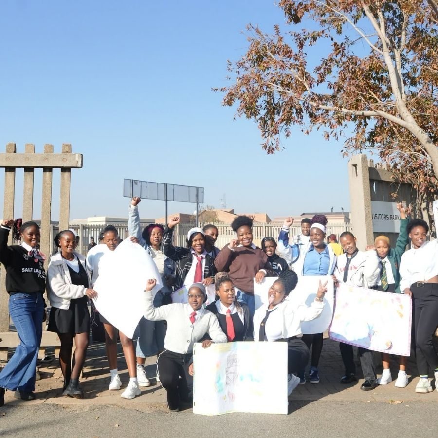 Ekurhuleni East TVET College students cheering with raised fists and posters outside a campus gate