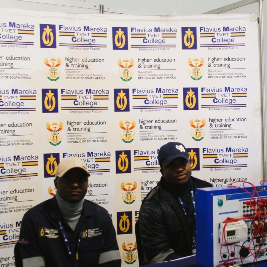 Flavius Mareka TVET College Students at Branded Backdrop Two Flavius Mareka TVET College students posing in front of a branded backdrop next to electrical training equipment.