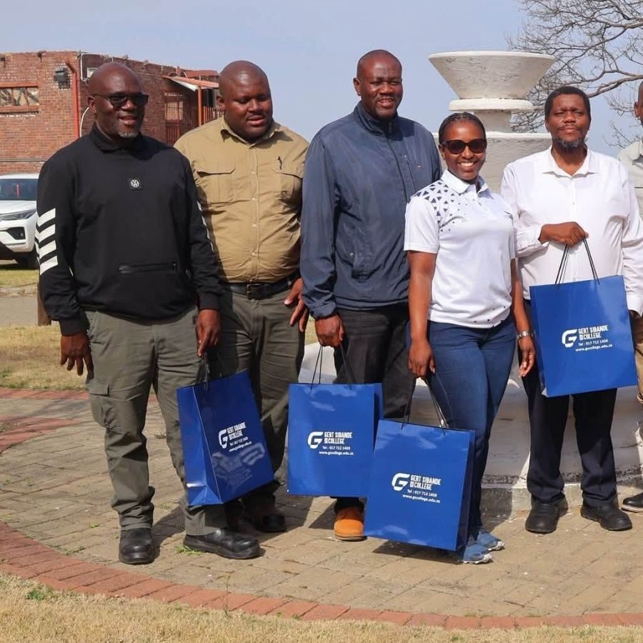 Gert Sibande TVET College Staff with Branded Gift Bags Five Gert Sibande TVET College staff members posing outdoors holding blue branded gift bags near a campus monument.