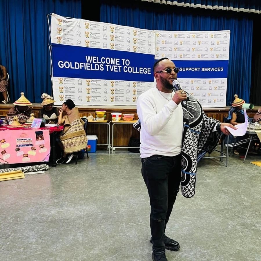 Speaker holding a microphone and traditional blanket on stage at a Goldfields TVET College Cultural Day celebration.