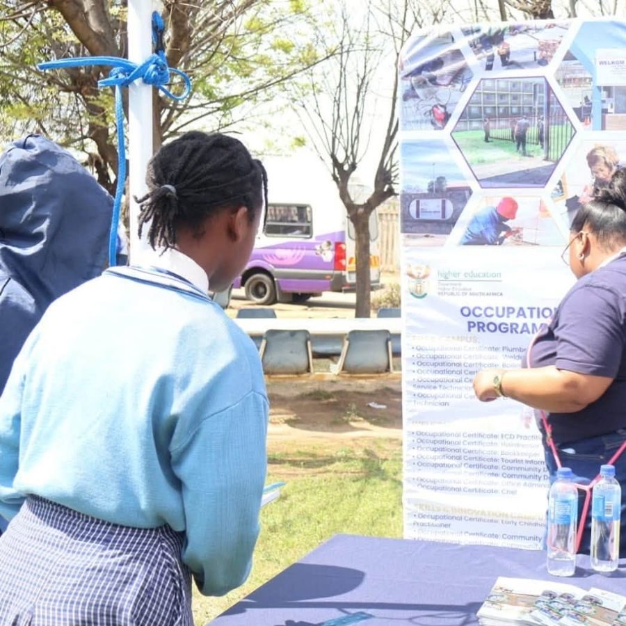 Goldfields TVET College student consults with a staff member beside an occupational programmes information banner outdoors.