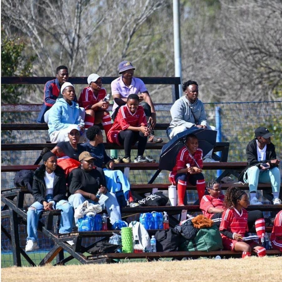 Majuba TVET College Supporters in Stadium Seating at Sports Day Majuba TVET College students and supporters, some in red team kit, sitting on outdoor stadium benches on a sunny day