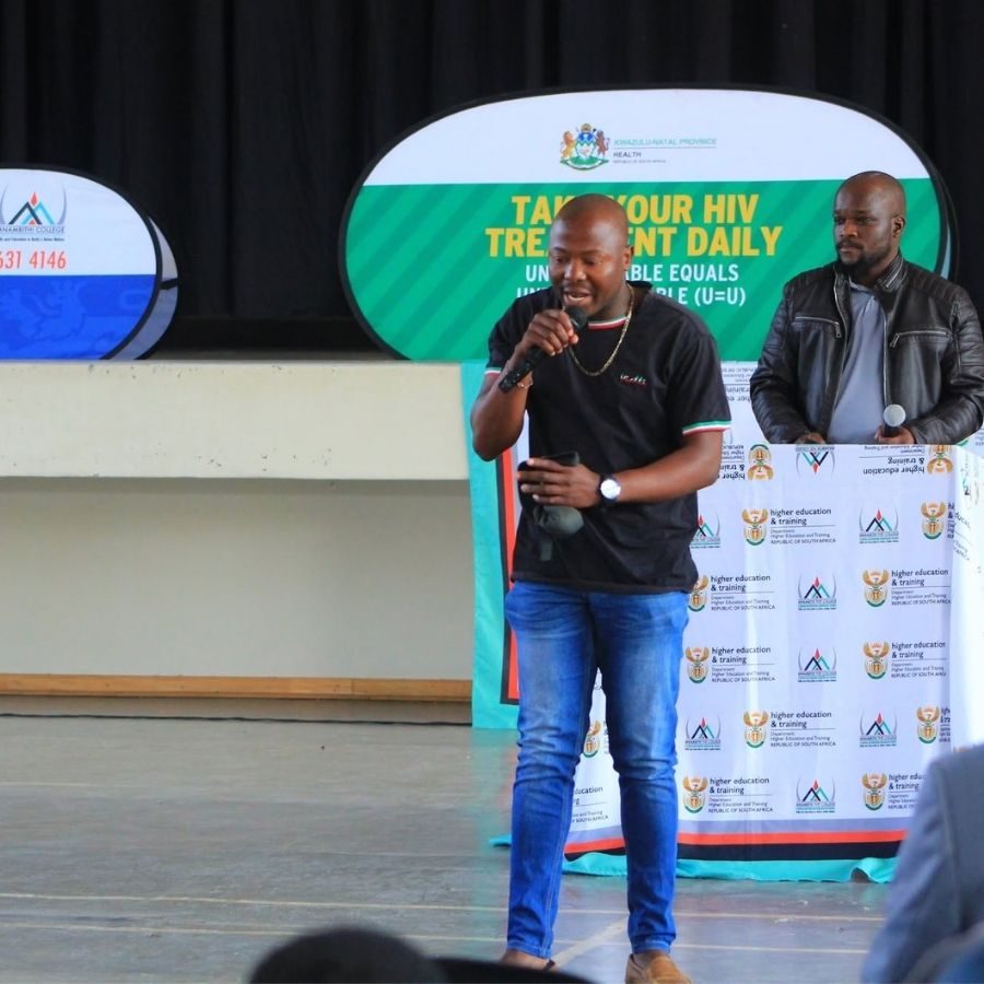 Mnambithi TVET College HIV Treatment Awareness Talk Speaker with microphone addressing students in front of a KwaZulu-Natal Health "Take Your HIV Treatment Daily" banner at Mnambithi College