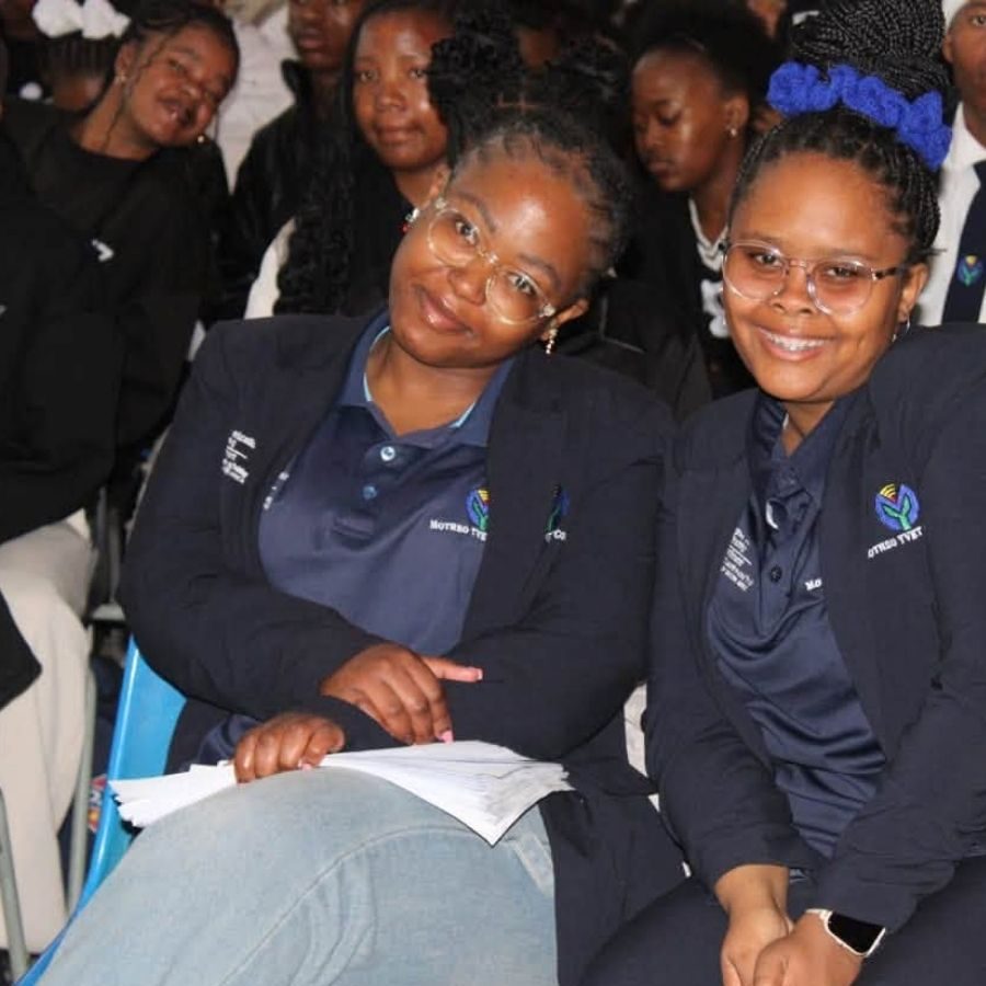 Two Motheo TVET College Students in College Uniform Two smiling Motheo TVET College students in navy blazers with the college logo, seated among classmates