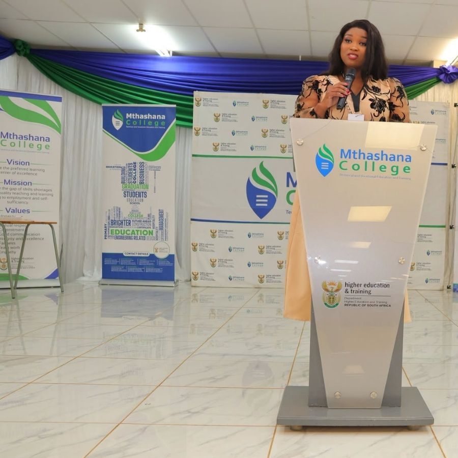 Speaker at Podium During Mthashana College Event Woman speaking at a branded Mthashana College podium in front of banners describing the college's vision and mission