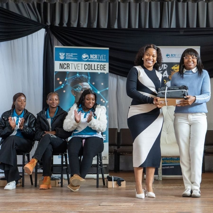 Staff exchange a gift box on stage at a Northern Cape Rural TVET College awards event, students in college shirts seated nearby.