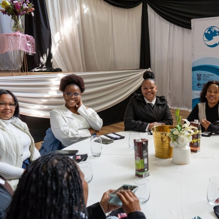 Women seated around a decorated round table smile for a photo at a Northern Cape Rural TVET College function.