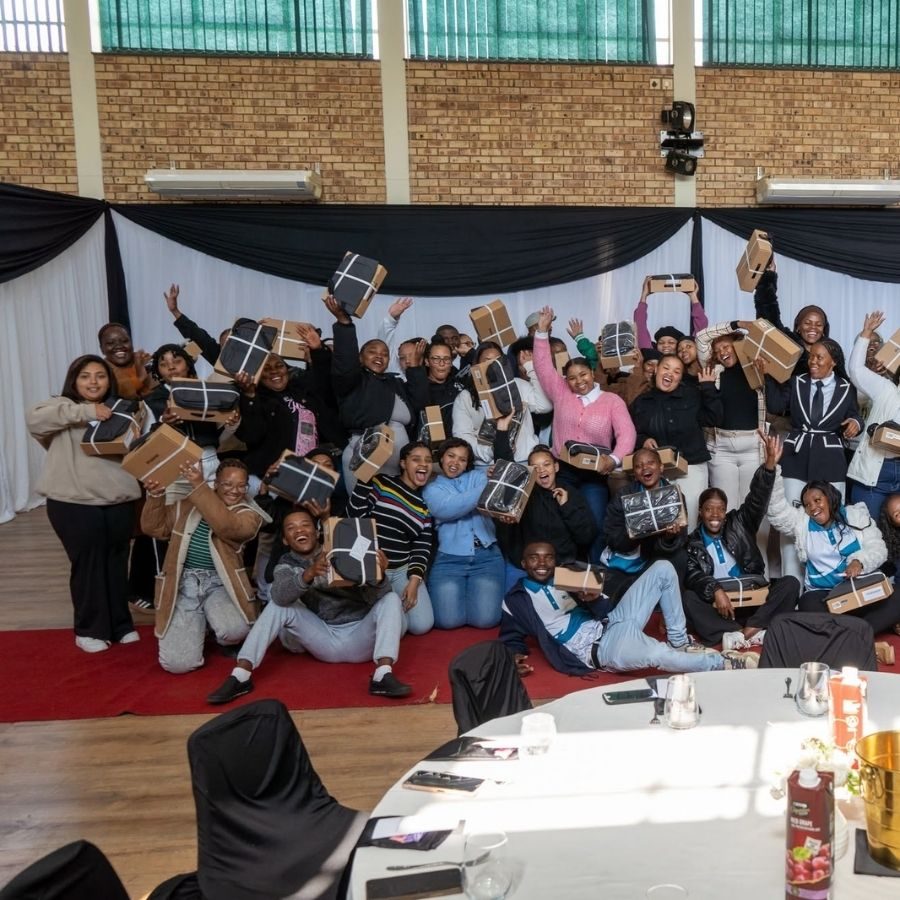 A large group of Northern Cape Rural TVET College students cheer and hold wrapped gift boxes in a packed hall.