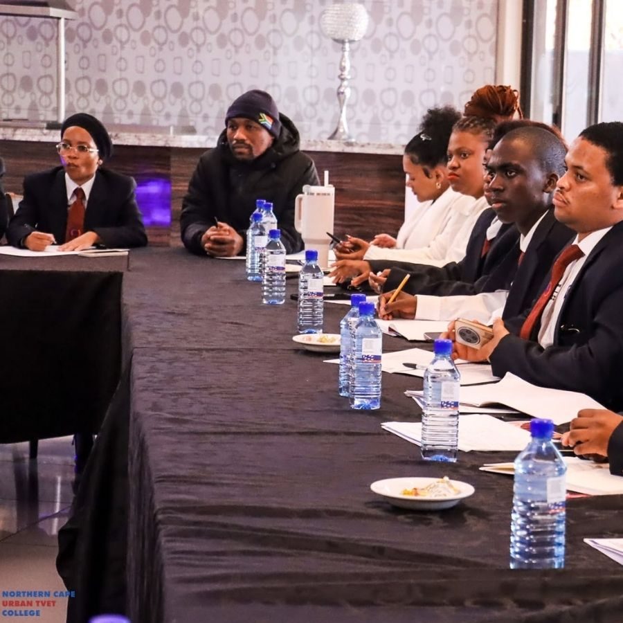 Staff and guests in formal attire sit around a long table during a Northern Cape Urban TVET College meeting.