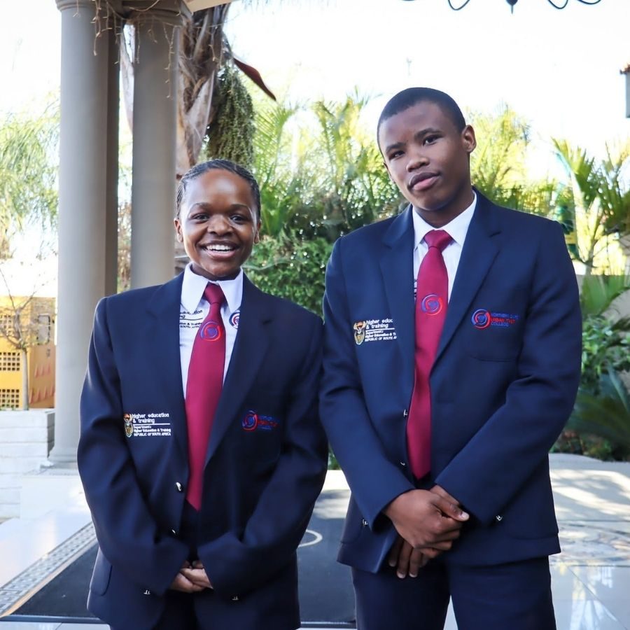 Two Northern Cape Urban TVET College students pose in navy blazers with red ties outside a building.