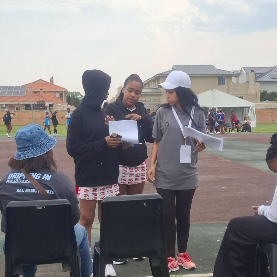 Officials review paperwork courtside at a Port Elizabeth TVET College sports day, athletes competing behind them.