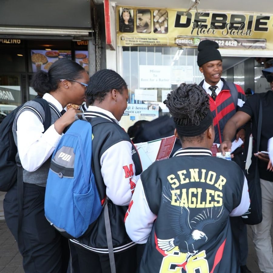 Tshwane North TVET College Recruiter Engages Learners in Town A Tshwane North TVET College recruiter in a black beanie hands out information to learners wearing varsity jackets outside shops