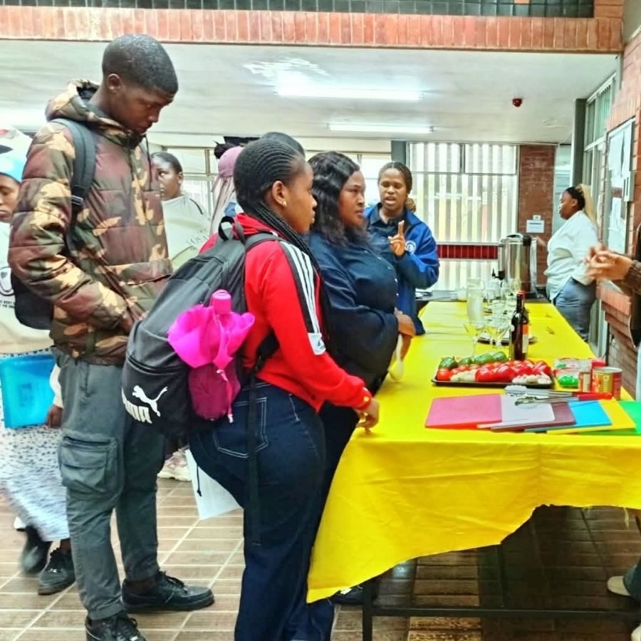 Umgungundlovu TVET College students gather around a table set with fresh produce and glassware during a hospitality demonstration