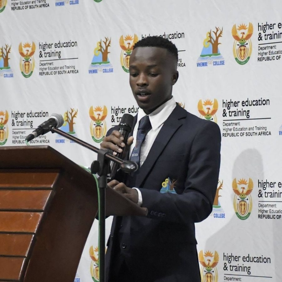 Vhembe TVET College Student Speaks at Podium A Vhembe TVET College student in a suit speaks into a microphone at a podium in front of the college step-and-repeat banner